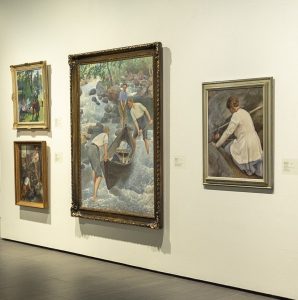 Kuva taidemuseon näyttelystä, jossa on neljä seinälle ripustettua maalausta. Keskimmäisenä on suuri maalaus, jossa kolme nuorta miestä vetävät soutuvenettä koskessa.