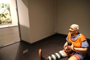 Vanhaa istuvaa naista esittävä veistos on sijoitettuna lattialle. Naisella on käsissään pieni apina.