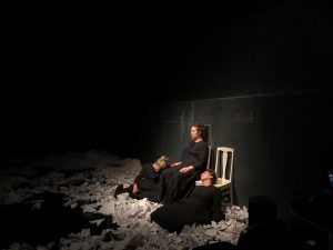 Kolme mustiin mekkoihin pukeutunutta naista, joista kksi istuu tuolilla kahden lattialla veltosti istuvan naisen välissä. Heidän ympärillään on rypisteltyjä papereita.