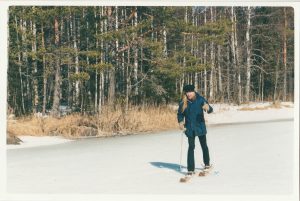 Pitkähiuksinen mies hiihtää hangella. Aurinko paistaa. Miehellä on päällään sininen talvitakki sekä housut, ja musta pipo.