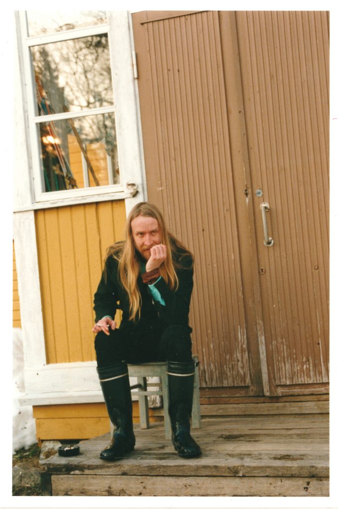 Pitkähiuksinen mies istuu keltaisen talon portailla. Hänellä on päällään mustat vaatteet ja kumisaappaat. Miehellä on mietiskelevä ilme, kyynärpää nojaa polveen ja hän pitelee leukaansa kädellään. Toisessa kädessä on tupakka.