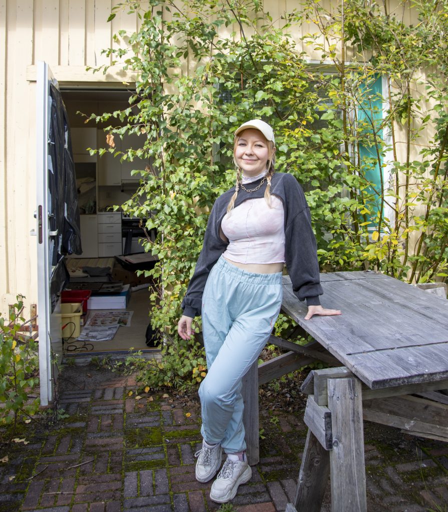 Lippalakkipäinen nainen nojailee jalat ristittynä pihalla olevaan pöytätasoon. Taustalla on avoin työhuoneen ovi, jonka vieressä rakennuksen peittää runsas kasvusto.