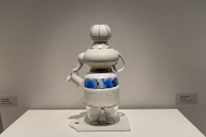 Posliiniastioita ja teekannuja asetettuna kahden lasin päälle tarjottimelle. Astioiden muoto vaikuttaa kannua kantavalta henkilöltä.