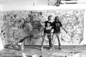 Kaksi miestä liikkuu suurikokoisen maalatun teoksen edessä. Miesten vaatteet ovat maalin tahrimat. Maaliin sotkeutuneella lattialla on maalipönttöjä ja muita teoksen tekemiseen liittyviä materiaaleja.