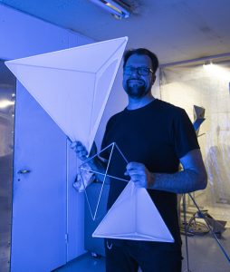 Parrakas t-paitaan pukeutunut mies pitelee kädessään läpinäkyvästä valkoisesta kankaasta ja metalliputkista rakennettuja pyramidimuotoja.