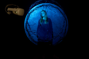 Nainen seisoo seinälle projisoidun kuulta vaikuttavan muodon edessä edessä. tilassa roikkuu pyöreä muovimateriaalista punottu kehä ja vr-lasit.