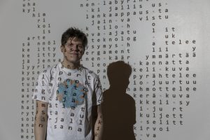 T-paitaan pukeutunut mies seisoo valkoisen seinän edessä, johon on projisoitu tekstiä. Projisointi osuu miehen päälle.