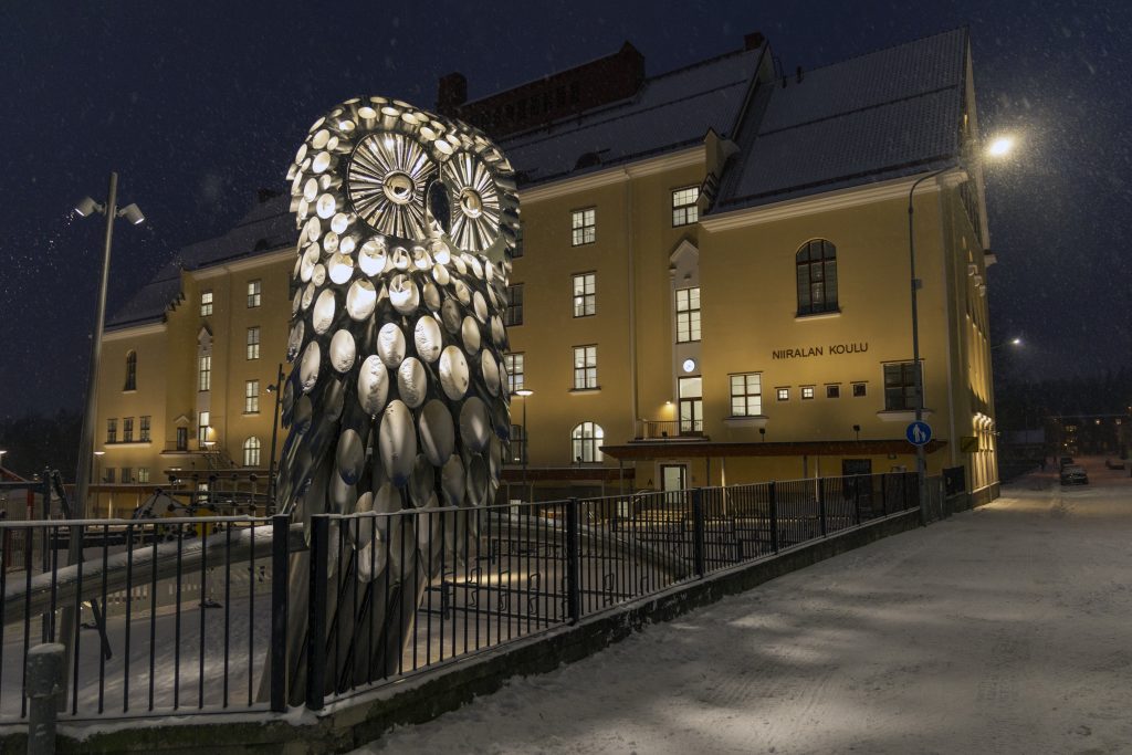Valaistu suurikokoinen pöllöveistos koulun pihan reunalla. Taustalla koulu.
