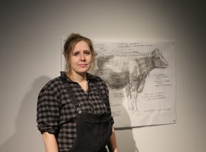 Ruutupaitainen nainen seisoo lehmää esittävän piirustuksen edessä. Piirustuksessa on nuolia ja tekstiä liittyen lehmän harjaamiseen.