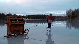Nainen luistelee märällä jäällä vetäen perässään vanhaa kelkkaa, jonka kyydissä on urkuharmooni.