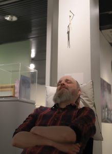 Harmaapartainen ruutupaitainen mies nojailee istualtaan tyynyyn teoksen alla silmät kiinni.