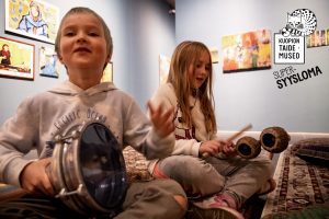 Kaksi lasta istuu taidemuseon näyttelytilan lattialla matolla ja soittaa vauhdikkaasti rytmisoittimia.