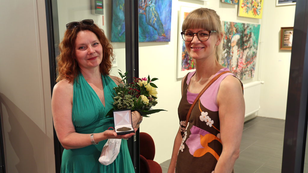 Kaksi naista, toisella kädessä kukkia ja mitali. Taustalla Taidelainaamon tilat taidekuvineen