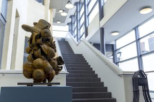 Kuvassa on keraaminen ruosteensävyinen veistos, jossa on kulhoja ja kumppareita kasassa. Taustalla on portaikko.
