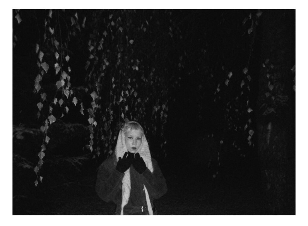 Mustavalkoinen kuva ulkotilasta pimeässä. Kuvan keskellä seisoo henkilö, jolla on huivi pään yli ja kädet nostettuna kasvojen eteen Henkilöllä on tummat käsineet puettuna. Taustalla näkyy puita, jotka muodostavat toistuvan kuvion pimeää taustaa vasten. Henkilö katsoo suoraan kameraan.