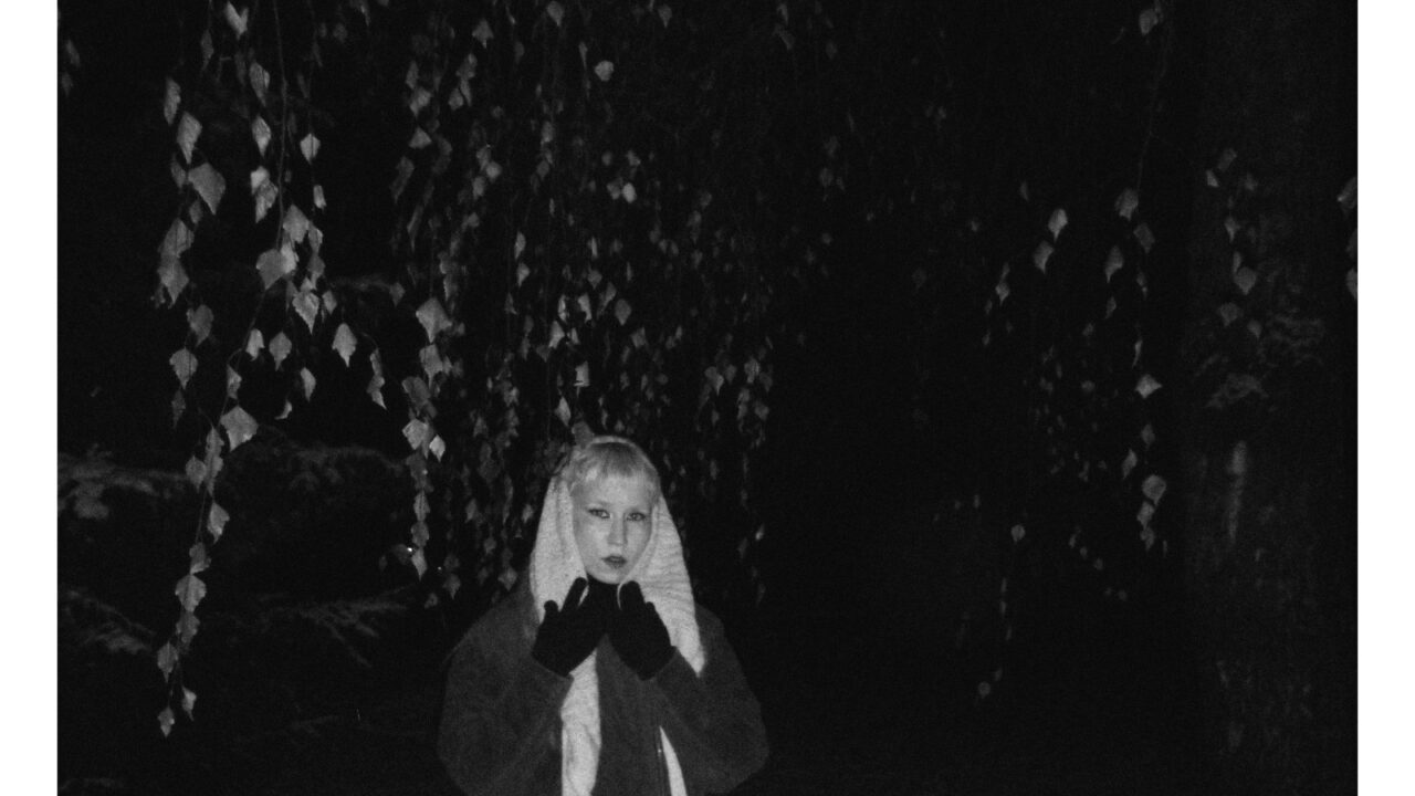 Mustavalkoinen kuva ulkotilasta pimeässä. Kuvan keskellä seisoo henkilö, jolla on huivi pään yli ja kädet nostettuna kasvojen eteen Henkilöllä on tummat käsineet puettuna. Taustalla näkyy puita, jotka muodostavat toistuvan kuvion pimeää taustaa vasten. Henkilö katsoo suoraan kameraan.