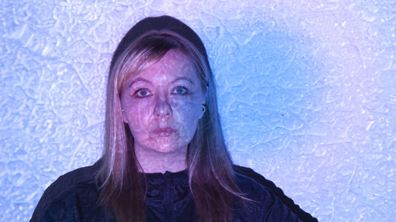 Henkilö kuvattuna edestä rintakuvan rajauksessa, tumma yläosa päällä. Kasvoille ja taustalle heijastuu videoprojisoinnin valoa sinisen ja violetin sävyissä, muodostaen epäsäännöllisen, tekstuurimaisen kuvion vaalealle seinälle. Hahmo katsoo suoraan kameraan.