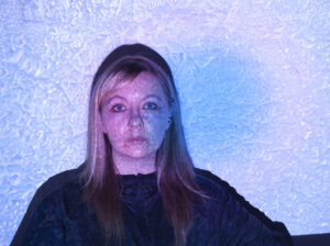 Henkilö kuvattuna edestä rintakuvan rajauksessa, tumma yläosa päällä. Kasvoille ja taustalle heijastuu videoprojisoinnin valoa sinisen ja violetin sävyissä, muodostaen epäsäännöllisen, tekstuurimaisen kuvion vaalealle seinälle. Hahmo katsoo suoraan kameraan.