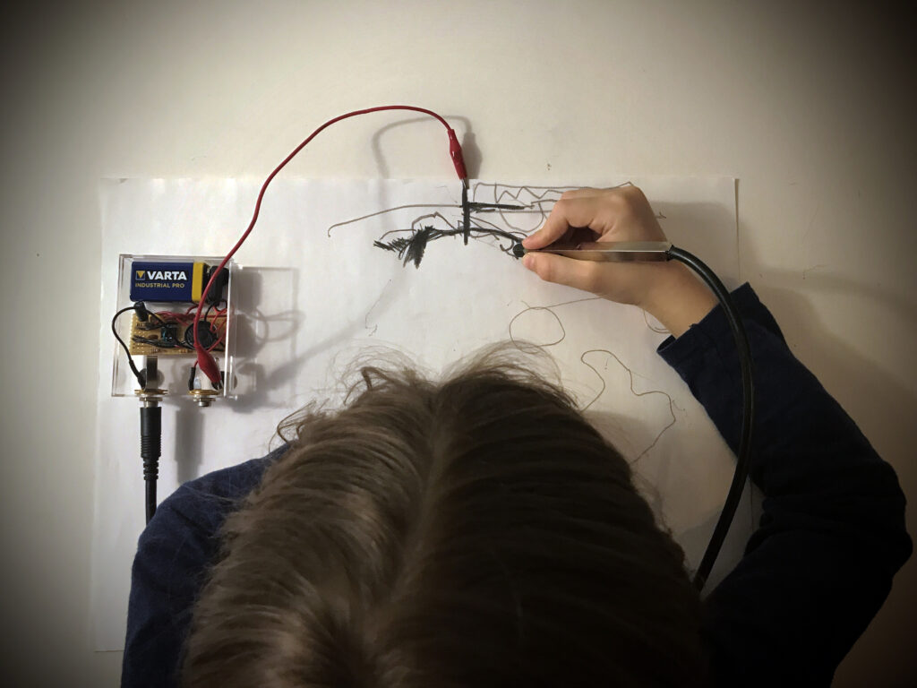 Henkilö piirtää sähköisellä kynällä paperille. Piirustus on kytketty johdoilla elektroniseen laitteeseen, joka tuottaa ääntä piirtämisen aikana. Kuvassa näkyy käsi, piirustuspaperi sekä ääntä tuottava elektroniikka.