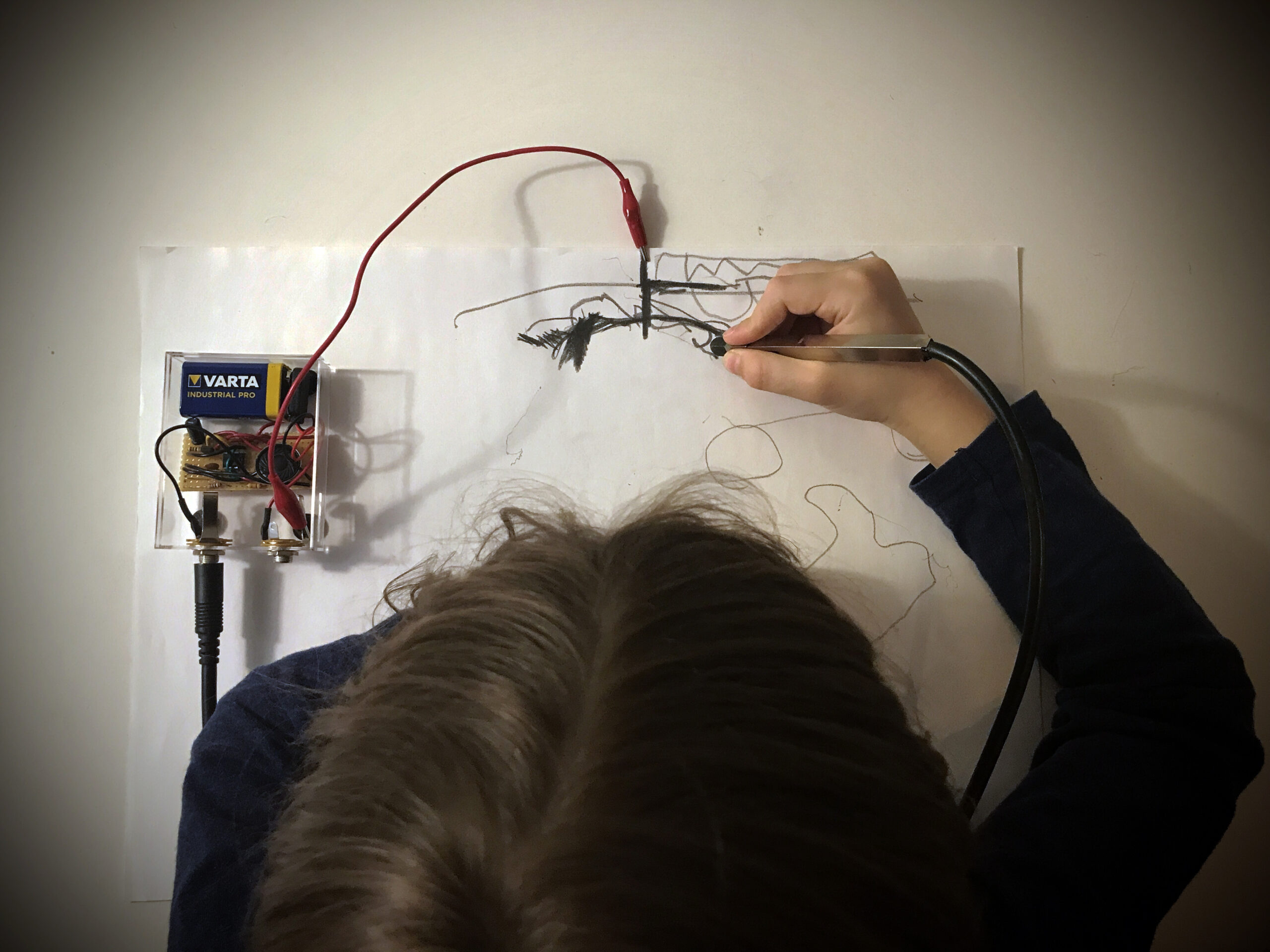 Henkilö piirtää sähköisellä kynällä paperille. Piirustus on kytketty johdoilla elektroniseen laitteeseen, joka tuottaa ääntä piirtämisen aikana. Kuvassa näkyy käsi, piirustuspaperi sekä ääntä tuottava elektroniikka.