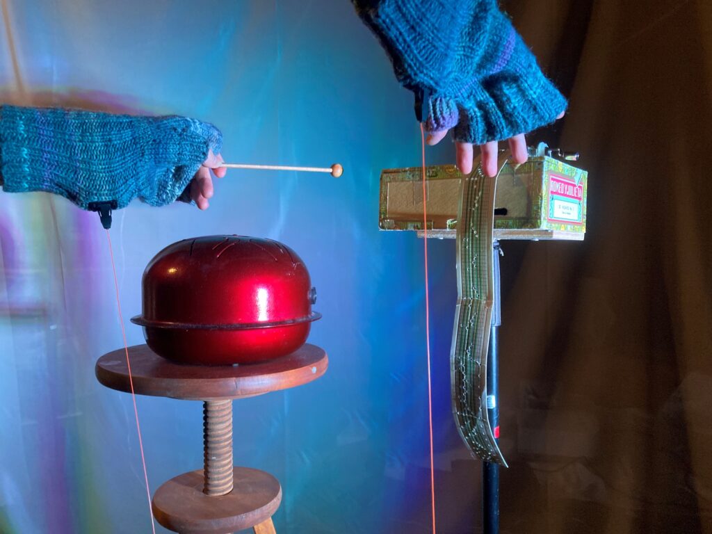 Neulotuilla käsineillä varustetut kädet käyttävät ääntä tuottavaa laitetta. Kuvassa näkyy punainen metallinen äänisoitin jalustalla sekä elektronisia osia sisältävä rakennelma, joiden avulla liike muuttuu ääneksi.
